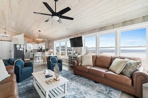 A spacious living area with a large window, a ceiling fan, and a kitchen in the background. - Salty Kisses & Starfish Wishes by Brunswick VR: Oceanfront & Pet-Friendly (Ocean Isle Beach)