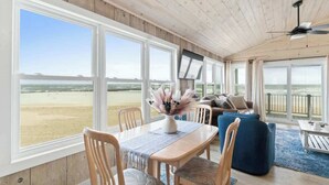 A room with a wooden ceiling, large windows with a beach view, a dining table with chairs, and a blue sofa. - Salty Kisses & Starfish Wishes by Brunswick VR: Oceanfront & Pet-Friendly (Ocean Isle Beach)