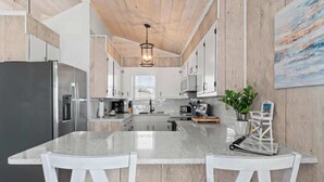 A modern kitchen with a central island, white cabinetry, and a wooden ceiling. - Salty Kisses & Starfish Wishes by Brunswick VR: Oceanfront & Pet-Friendly (Ocean Isle Beach)