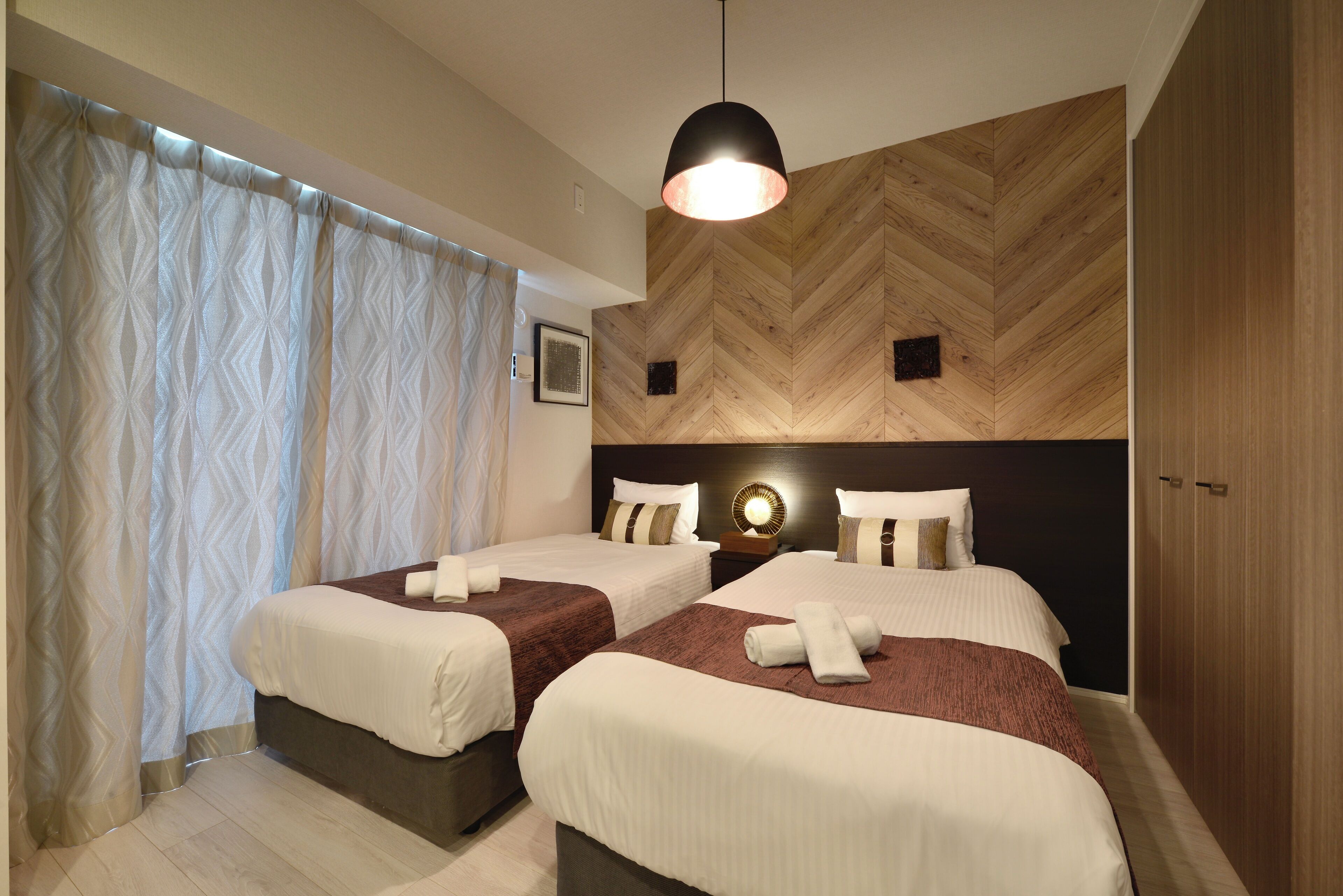 A hotel room with two beds, a wooden herringbone wall, a hanging light fixture, and a large wardrobe.