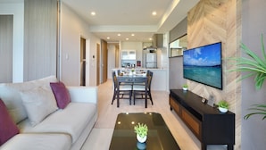 A modern living room with a sofa, a coffee table with a plant, a dining area with a table and chairs, and a kitchen in the background. - Branchera naha akebono premist R69 (Naha)
