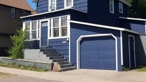 A two-story house with a blue exterior and white trim, a front porch, and a garage. - Adventure House: Fully Stocked Kitchen 5 mins from Michigan Tech (Hancock)