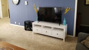 A living room with a flat-screen TV on a white TV stand, a clock on the wall, and two vases with yellow flowers. - Adventure House: Fully Stocked Kitchen 5 mins from Michigan Tech (Hancock)