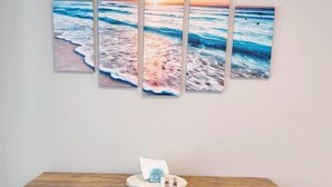 Un coin repas avec une table et des bancs en bois, une œuvre d’art représentant une scène de plage accrochée au mur, et un petit objet blanc posé sur la table.