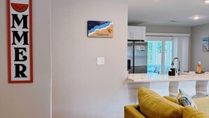 Un salon moderne avec un canapé jaune, une cuisine en arrière-plan et une pancarte « bonjour, été » accrochée au mur.