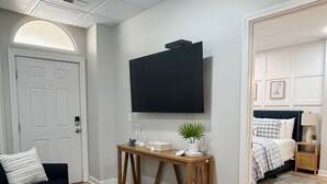 A modern living room with a flat-screen TV mounted on the wall, a dark-colored sofa, a wooden console table, and a patterned rug. - ☀️Beachfront☀️ 2Br 2Ba with Heated Pool, Playground, Gym, and Arcade! (North Topsail Beach)