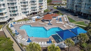 A hotel with a large pool surrounded by lounge chairs and umbrellas, multiple buildings with balconies, and a parking area. - ☀️Beachfront☀️ 2Br 2Ba with Heated Pool, Playground, Gym, and Arcade! (North Topsail Beach)