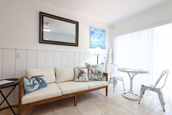 A living room with a sofa, a small table, a chair, a glass-top table, a lamp, a painting, and a large mirror on the wall. - Beach Tree across street from beach (Siesta Key)