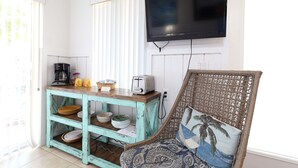 Una cocina con una mesa de madera color aguamarina, un televisor de pantalla plana negro montado en la pared y una silla de mimbre con un cojín estampado.
