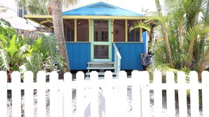 A small, single-story building with a blue roof and yellow walls, surrounded by a white picket fence and palm trees. - Blue Dolphin Cottage steps from the beach (Siesta Key)