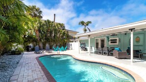 A swimming pool with a brick border, surrounded by palm trees and a covered patio area with outdoor furniture.