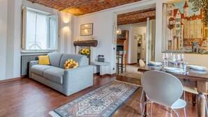 Een woonkamer met een bank, een eettafel met stoelen, een kamerkleed met patroon en een bakstenen plafond.