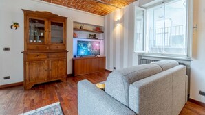 Een woonkamer met een bakstenen plafond, een houten kast, een televisie, een grijze bank en een raam met uitzicht.