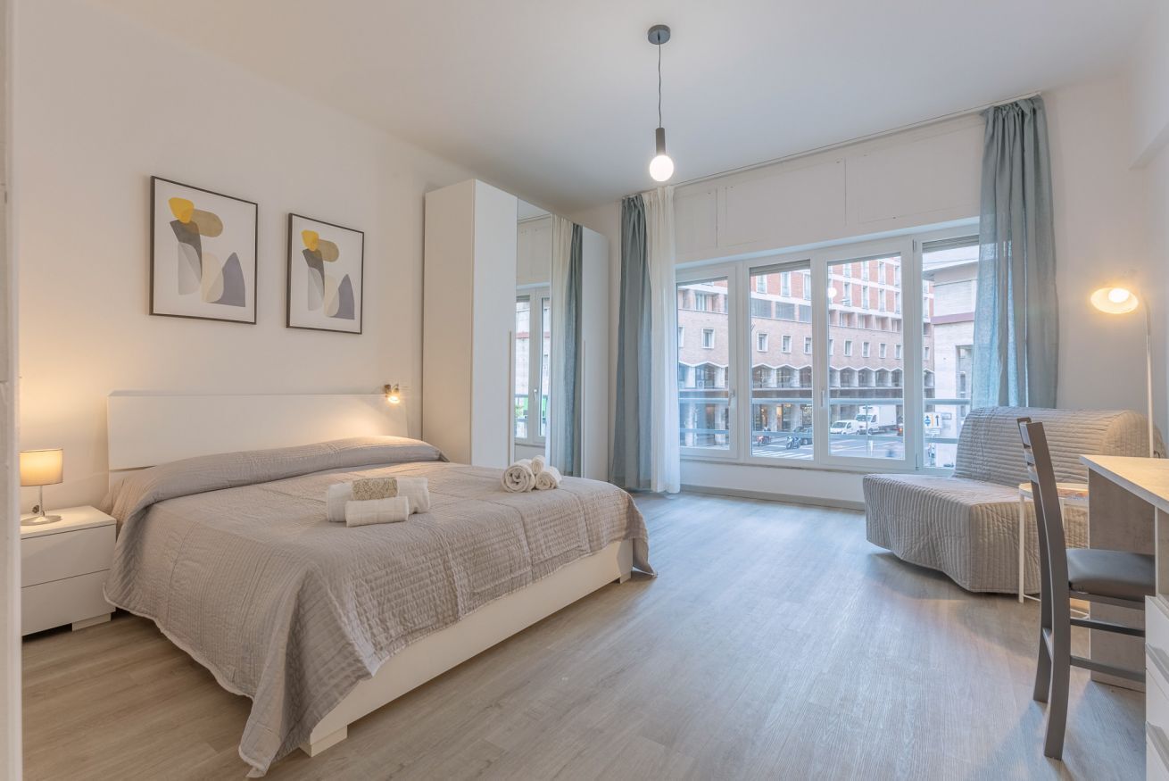 A modern bedroom with a large bed, bedside tables, a desk, and a view of the city through large windows.
