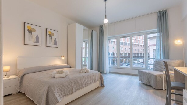 Ein modernes Schlafzimmer mit einem großen Bett, Nachttischen, einem Schreibtisch und Blick auf die Stadt durch große Fenster.