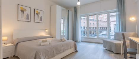 A modern bedroom with a large bed, bedside tables, a desk, and a view of the city through large windows.