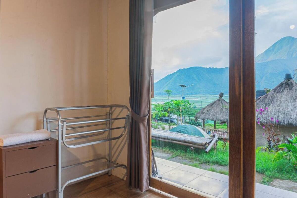 Une chambre avec un lit en métal, une table de chevet en bois et une vue sur un paysage tropical, avec des montagnes et des constructions aux toits de chaume.