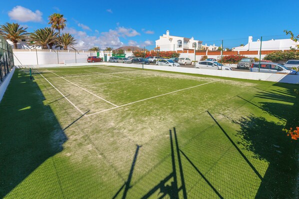 Un court de tennis avec un filet, entouré d’une clôture blanche, avec des voitures garées et des bâtiments en arrière-plan.