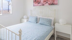 Ett sovrum med en säng, vita sängbord, en vit sänggavel, ett fönster med utsikt och ett blommigt konstverk som hänger på väggen.