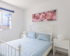 Una camera da letto con un letto, comodini bianchi, una testata del letto bianca, una finestra vista e un quadro floreale appeso al muro.