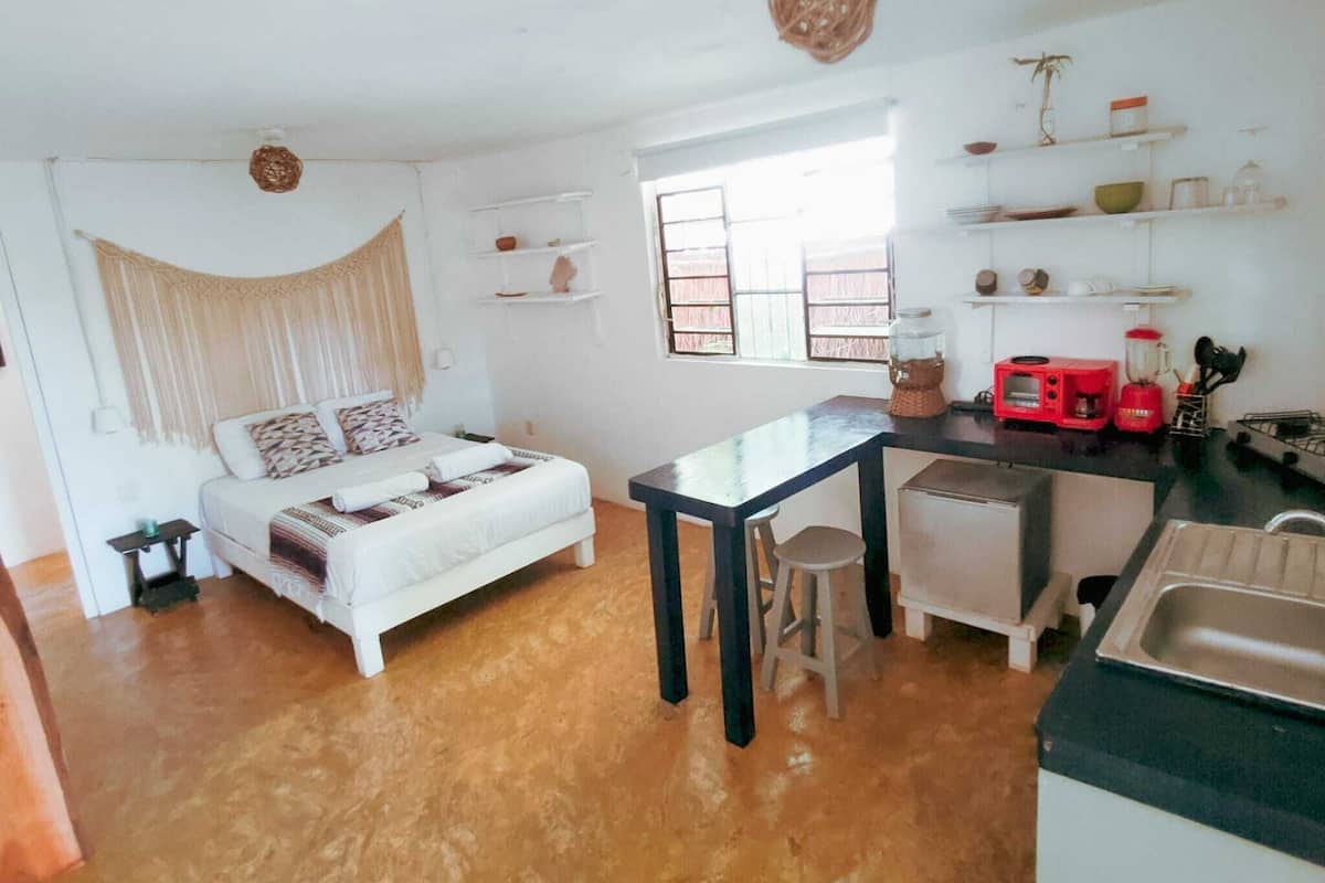Una habitación compacta con cama, una mesita, un taburete, kitchenette y ventana.