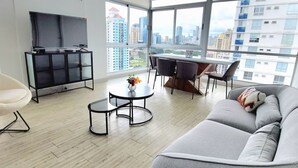 Ein modernes Wohnzimmer mit einem grauen Sofa, einem runden Couchtisch, einem Fernseher auf einem schwarzen Standfuß und einem Essbereich mit Tisch und Stühlen.