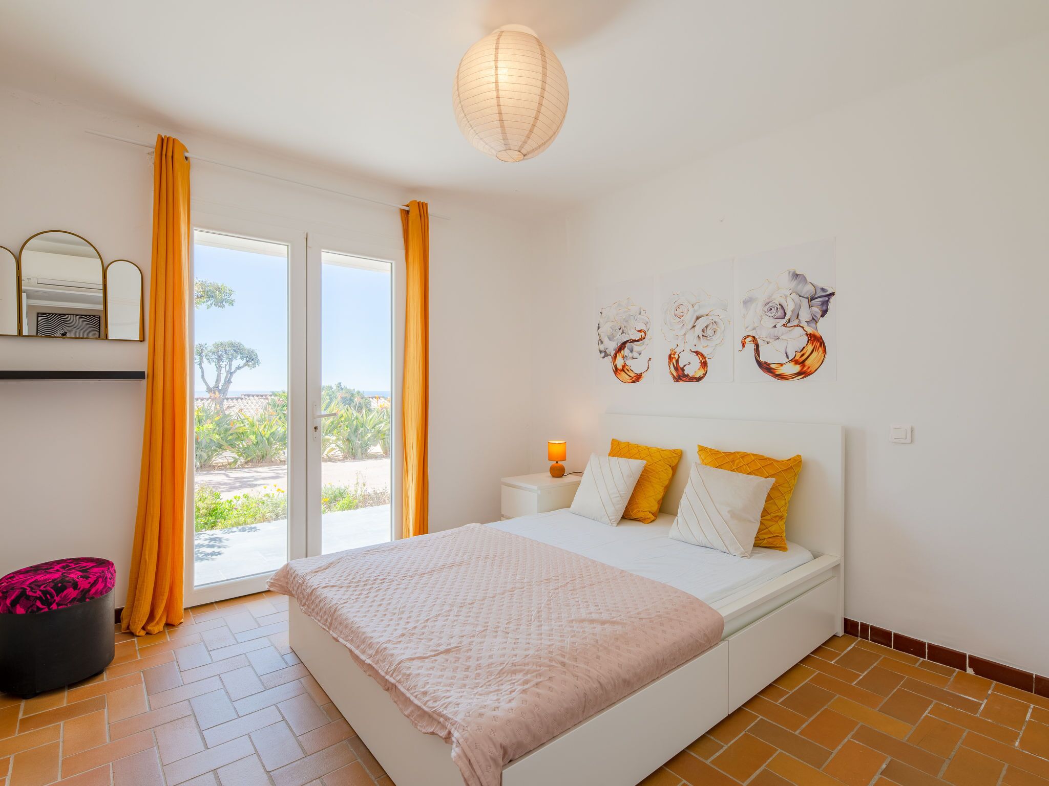A bedroom with a bed, a nightstand, a lamp, a window with curtains, and a view of greenery outside.