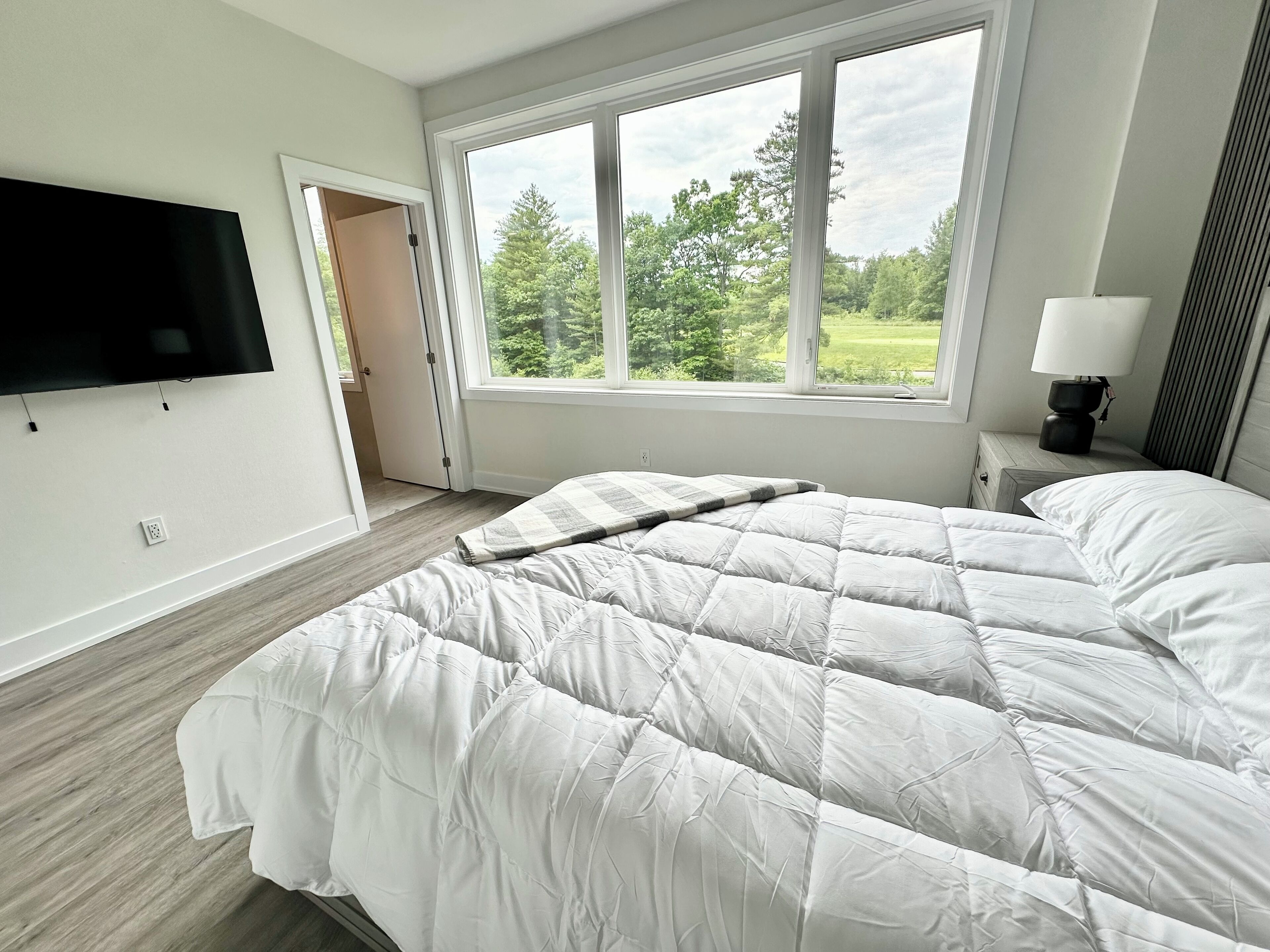 Een slaapkamer met een groot bed, een aan de muur bevestigde televisie, een raam met uitzicht op bomen, en een nachtkastje met een lamp.