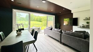 A modern living room with a dining area, large sliding glass doors to a garden, a flat-screen TV mounted on the wall, and a wooden ceiling.