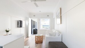 Une chambre moderne et minimaliste, dotée d’un téléviseur à écran plat, d’une table d’appoint en osier, d’un canapé blanc et offrant une vue sur l’extérieur par une fenêtre.