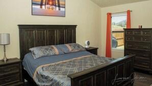 A bedroom with a wooden bed, a nightstand with a lamp, a dresser, and a window with curtains. - Cozy 4BR Retreat Near Randolph AFB & Ft Sam (Converse)