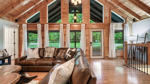 A spacious living room with a brown leather sectional sofa, a wooden floor, and large windows offering a view of greenery outside. - 6BR-Epic Yard-Game And Movie Rooms (Sevierville)