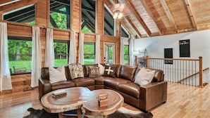 A cozy living room with a large L-shaped sectional sofa, a round wooden coffee table, and a view of a lush green landscape through large windows. - 6BR-Epic Yard-Game And Movie Rooms (Sevierville)