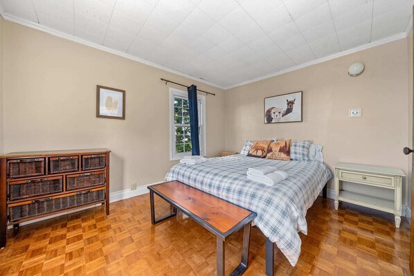 A bedroom with a bed, a wooden dresser, a bench, a nightstand, and a window with curtains.