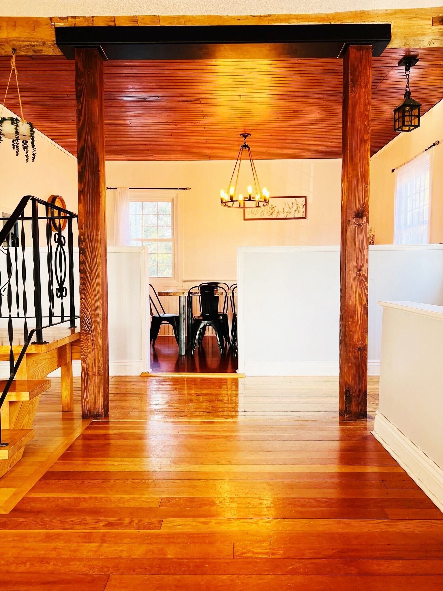 A spacious room with wooden flooring, a dining area with chairs, a chandelier, and a staircase with a railing.