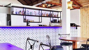 A bar area with a tiled backsplash, a wooden ceiling, and a bar counter with shelves of bottles. There are metal chairs and a round table in the foreground.