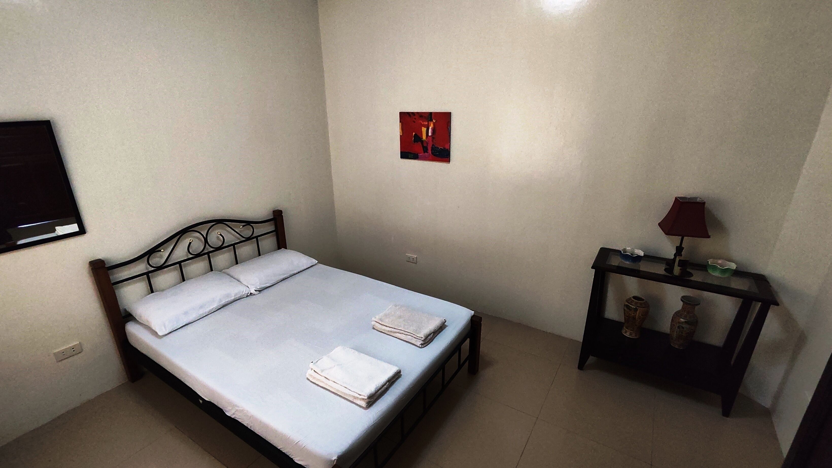 A bedroom with a bed, a bedside table with a lamp, and a small painting on the wall.