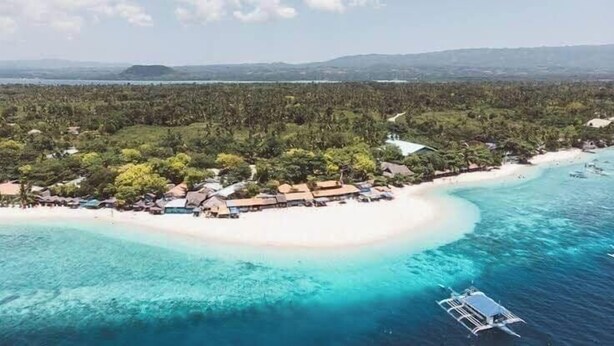 An aerial view of a coastal resort with a white sandy beach, clear turquoise waters, and a dense forested area.