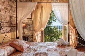 A bedroom with a stone wall, a bed with floral bedding, a view of greenery, and a balcony with metal railings. - Holiday Home 'Villa Silvia' with Private Pool, Wi-Fi and Air Conditioning (Rignano sull'Arno)