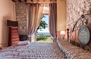 Ein Schlafzimmer mit Bett, Kommode, Fernseher, Lampe und Blick ins Freie.
