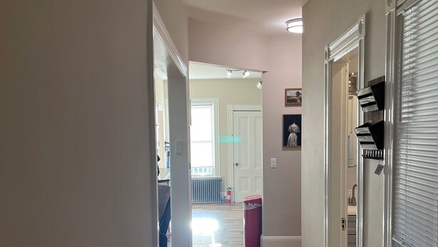 Un passadís amb sòl de fusta, parets blanques i una porta que dona a una habitació amb un radiador i un gresol.