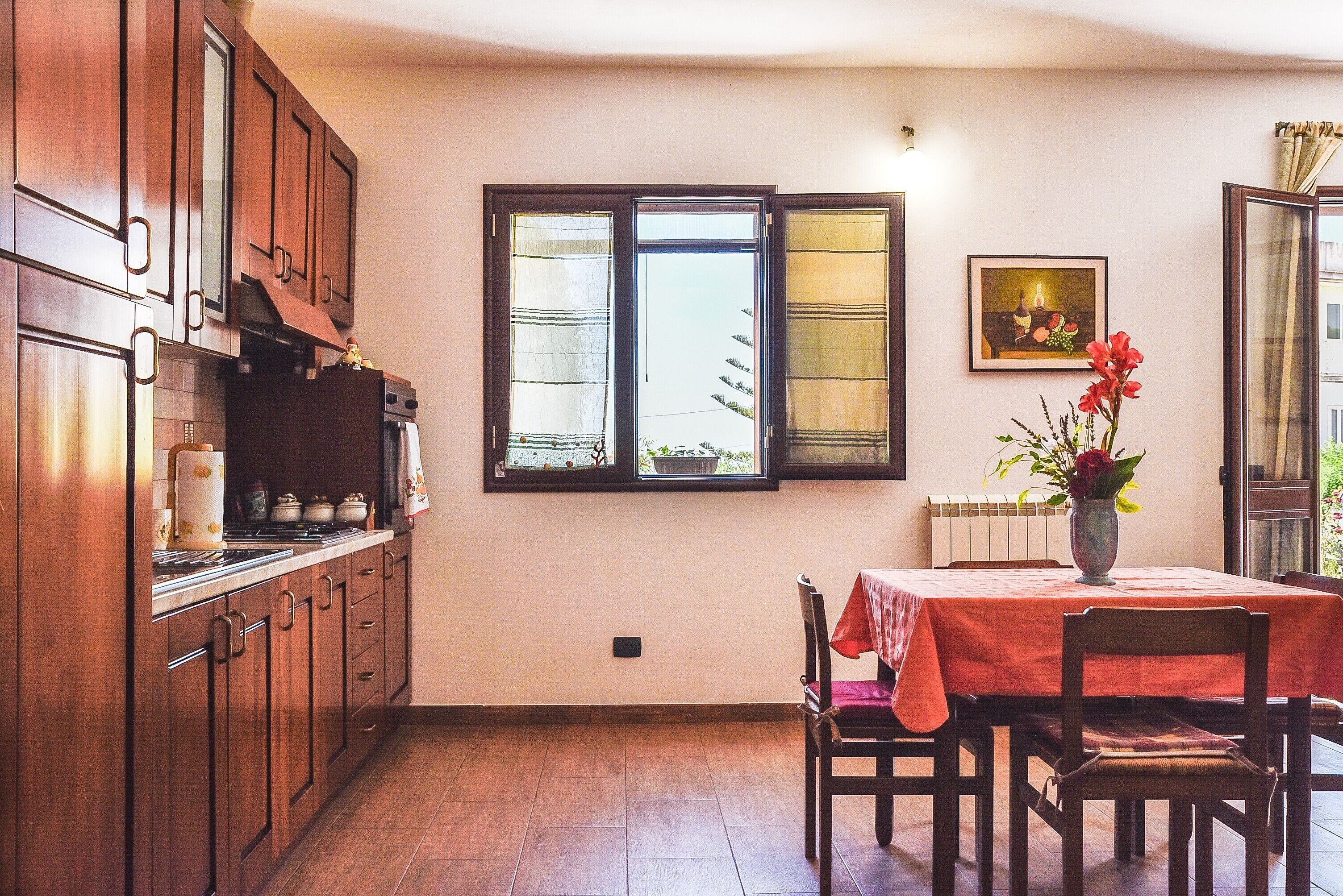 A kitchen with wooden cabinets, a dining table with chairs, a window with curtains, and a vase with flowers.