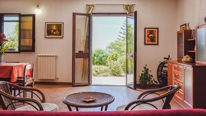 Une pièce comprenant une table à manger, des chaises, une petite table ronde, un radiateur, un meuble et une grande porte-fenêtre coulissante donnant sur un espace extérieur verdoyant.