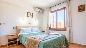 Quarto com cama, mesa de cabeceira, ar condicionado fixado na parede, janela com cortinas e luminária na parede.