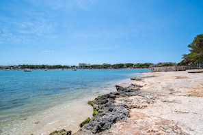 A coastal beach with clear water, rocky shoreline, and distant buildings.
