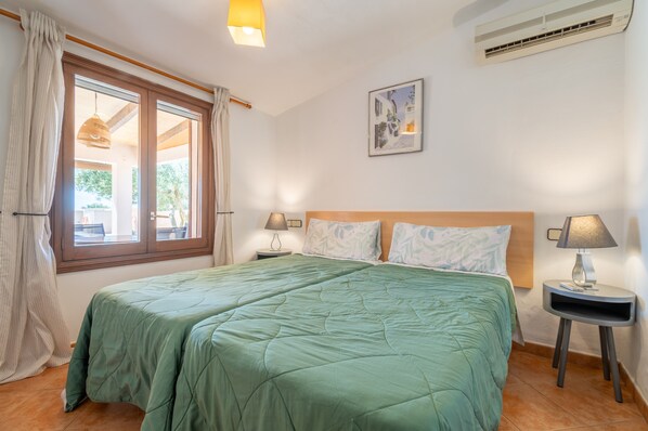 A double bed with a green bedspread, two bedside tables with lamps, a window with curtains, a wall-mounted air conditioner, and a framed picture. - Villa 'Capricho' with Private Pool, Wi-Fi, and Air Conditioning (Illes Balears)