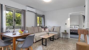 Una sala moderna con sofá, mesa de centro y mesa de comedor. Hay ventanas con cortinas, una mesita auxiliar y una planta.