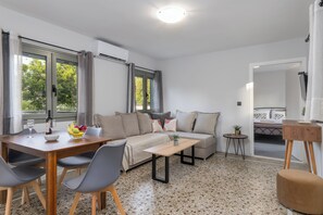 A modern living room with a sofa, coffee table, and dining table. There are windows with curtains, a small side table, and a plant.