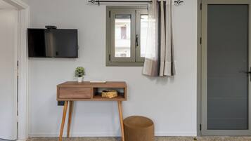 Une pièce avec un téléviseur fixé au mur, un petit bureau en bois avec un panier, un tabouret rond et une porte avec une fenêtre.