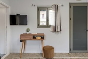 A room with a TV mounted on the wall, a small wooden desk with a basket, a round stool, and a door with a window.